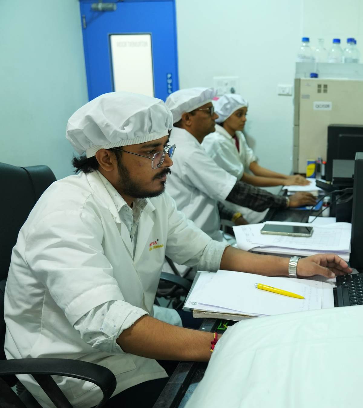 Technical staff in white coats and hair caps working at computers with documentation in the facility